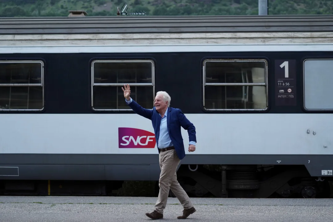 Un homme en veste bleue court et salue de la main le long d'un train.